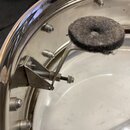 Detailní pohled na vnitřek bubnu s kovovým obručím a bílou blánou. Na obruči je připevněno kovové rameno s kulatým tlumicím polštářkem z šedého plsti…
