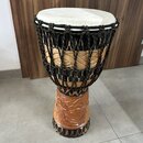 Djembe s bílou membránou a dřevěným korpusem. Nástroj je opleten černými provazy s barevnými detaily a spodní část korpusu obsahuje vyřezávaný orname…