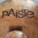 Detailní záběr na bronzový činel s koncentrickým žebrováním. Na povrchu je černý nápis pAiste a pod ním menší text 20" RIDE. Vpravo je vyražen nápis…