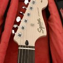 Hlava světlé elektrické kytary s hnedým hmatníkem, umístěná v červeném obalu. Na hlavě je černý nápis Squier by Fender a modelové označení JAZZMASTER…