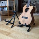 Dřevěný akustický ukulele s výřezem v těle, umístěné na černém stojanu. Vedle něj stojí druhý volný černý stojan. Na těle nástroje není vidět žádné l…