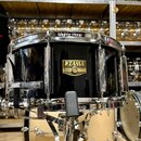 Černý malý buben s chromovaným kováním a obručemi. Na přední straně je zlatý štítek s nápisy TAMA, SNARE DRUM a SERIAL No. (číslo je nečitelné). Na h…