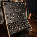 Tři černé syntezátory značky moog umístěné v dřevěném stojanu. Horní modul nese nápis SPECTRAVOX | SEMI-MODULAR ANALOG SYNTHESIZER, prostřední DFAM |…