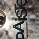 Detailní záběr na kovový povrch činely s viditelným broušením. Na povrchu je černý nápis Paiste a menší svislý text MADE IN SWITZERLAND. V horní část…