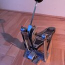 Kovový basový pedál s černým šlapadlem a bílo-černým beaterem. Na bočním kole jsou vidět nápisy "SUPERGLIDE" a "CHAIN DRIVE". Povrch kovových částí v…