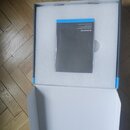 Otevřená bílá krabice s vložkou z bílého polystyrenu. Uvnitř leží šedý obálkový dokument s modrým horním okrajem. Na dokumentu je bílý nápis SENNHEIS…