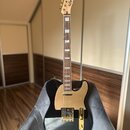 Černá elektrická kytara s tónem a ladicími kolíky v zlatém provedení. Na hlavě je nápis Squier TELECASTER. Kytara má zlatý pickguard a hриф s bílými…