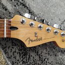 Fotografie zachicuje hlavu elektrické kytary z přírodního světlého dřeva s hnedým hmatníkem. Na hlavě je černý nápis Fender a STRATOCASTER®, v pravé…