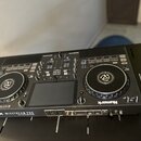 Černý DJ 컨트롤러 s dvěma jogovými koly a centrálním displejem. Na přední straně je bílý nápis Numark MIXSTREAM PRO a text v menším písmu pod ním. Na pra…
