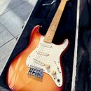 Elektrická kytara typu Stratocaster s lakovaným tělem v červeno-žlutém sunburst provedení a bílým pickguardem. Nástroj má javorový hmatník a tři sním…