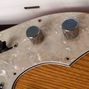 Detail elektrické kytary s bílým perleťovým pickguardem a dřevěným tělem s viditelnou masivní strukturou. Na pickguardu jsou umístěny dva stříbrné ko…