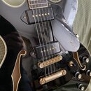 Černá polodupatá elektrická kytara s bílým lemováním a zlatým hardwarem. Nástroj má dva humbuckery, zlatý most a zlatý strunník. Na těle jsou tři čer…