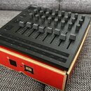 Černý MIDI kontrolér s řadou posuvníků a mřížkou knoflíků, umístěný na červeném kartonovém obalu. Na levé straně jsou viditelné nápisy „Dim Control“…