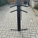 Černý kovový stojan na hudební nástroje s dvěma páry rozkládacích ramen. Stojan stojí na dlažbové ploše před garážovými vraty. Na povrchu nejsou vidi…