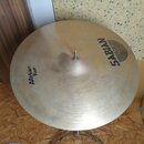 Zlatavý kovový činel na stojanu. Na povrchu je černý nápis SABIAN s doprovodným logem a text AA Heavy Ride. Povrch vykazuje jemné koncentrické rýhy a…