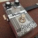 Kytarový efekt pedál značky ModTone s názvem COLISEUM Reverb a označením MT-RV. Stříbrný kovový šasi s černými lineárními vzory a dvěma ovládacími kn…