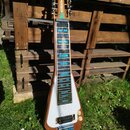 Elektrická lap steel kytara s dřevěným korpusem a bílým krycí plátem. Na hlavě je červený nápis Sondley a pod ním zelený nápis Steel Guitar. Hmatník…