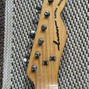 Hlava elektrické kytary z přírodního dřeva s šesti kovovými ladičkami. Na povrchu je černý nápis ELECTRIC GUITAR a pod ním stylizované logo luxor. Po…