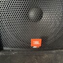 Detailní pohled na černý reproduktor s kovovou mřížkou z perforovaného plechu. Na spodní části mřížky je oranžový čtvercový štítek s bílým nápisem JB…