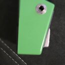 Detailní záběr na spodní část zeleného hudebního nástroje nebo příslušenství. Je vidět kovový vstup pro jack kabel. Povrch je hladký a bez viditelnýc…