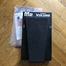 Černý volume pedál s textem M2 PASSIVE VOLUME. Na bocích jsou viditelné nápisy OUT a IN. Vedle pedálu je v plastovém sáčku dokumentace s nápisem MORL…