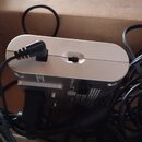 Stříbrný elektronický přístroj s obloukovanými hranami, do kterého je zapojen černý napájecí kabel. Přístroj leží v kartonové krabici s nápisem BATA…