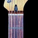 Fotografie zachycuje hmatník a hlavu elektrické kytary s javorovou hlavou a tmavým hmatníkem. Na hlavě je černé logo Fender a nápis STRATOCASTER®. V…