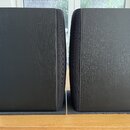 Dva černé reproduktory s povrchem imitujícím strukturu dřeva. Jsou umístěny vedle sebe na šedých podložkách z pěny. Na viditelných stranách nejsou žá…