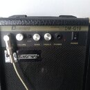 Kytarový zesilovač s černým povrchem z texturovaného materiálu a kovovým ovládacím panelem. Na panelu jsou nápisy GUITAR AMPLIFIER 10WATTS DM-G10 a l…