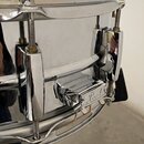 Detailní pohled na chromovaný snare drum. Na mechanismu napínání je viditelné logo Pearl. Povrch je lesklý a v dobrém stavu, s viditelnými odlesky ok…