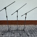 Tři šedé mikrofonní stojany s rameny. Stojany mají trojnožku a teleskopické části s černými utahovacími knoflíky. Povrch je matný, bez viditelných zn…