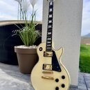 Krémová elektrická kytara typu Les Paul se zlatým hardwarem a černým pickguardem. Na hlavě hmatníku je viditelné logo Gibson. Nástroj má dva humbucke…