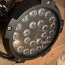 Černý kulatý LED reflektor s kovovým tělem a ochranným sklem. Pod sklem je vidět uspořádání více okruhů LED diod. Na horní části těla jsou vidět čtyř…