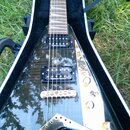 Černá elektrická kytara s trojúhelníkovým tělem v otevřeném kufru. Na obou snímačích je bílý nápis Seymour Duncan. Kytara má zlatý hardware, včetně m…