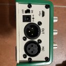 Záběr na zadní panel zařízení v zeleném kovovém rámu s bílým podkladem. Jsou zde vidět dva XLR konektory s nápisy OUT a MIC, USB port, napájecí vstup…