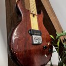 Elektrická kytara s tmavě hnědým dřevěným korpusem a světlým javorovým hmatníkem. Nástroj má jeden černý humbucker u kobylky, stříbrný strunník a dva…