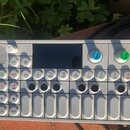 Stříbrný elektronický sekvencer s displejem, mluvčíkem a řadou tlačítek. Na ovládacích prvcích jsou viditelné symboly, čísla 1 až 5 a nápisy IN, OUT,…