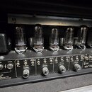 Fotografie zachycuje vnitřek kytarového zesilovače s řadou šesti elektronických lamp. Na spodním panelu jsou vidět konektory pro reproduktory s nápis…