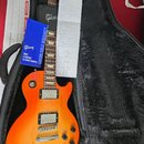 Elektrická kytara s žlutě-oranžovým gradientem v černém kufru. Na hlavě hmatníku je bílé logo Gibson a na modrém záručním listu nápisy Gibson, ONLY A…