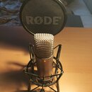 Kondenzátorový mikrofon v černém šokabsorbujícím držáku. Mikrofon má stříbrnou kovovou mřížku a tmavý trup s malým oranžovým prvkem. V pozadí je umís…