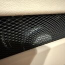 Detailní záběr na část reproduktoru s bílým povrchem z texturovaného materiálu. Střed fotky tvoří černá kovová mřížka s kosočtvercovým pletivem. Za m…