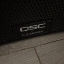 Detailní záběr na spodní část černého reproduktoru s kovovou mřížkou. Na čelním panelu je umístěn štítek s bílým logem QSC a nápisem K.2 SERIES. Povr…