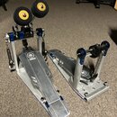 Stříbrný dvojpedál k basové bubnu s modrými detaily. Na obou pedálech je viditelné logo a nápis YAMAHA. Pedál má dvě černé beatery s žlutými středový…