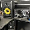 Na fotografii jsou tři studiové monitory a jeden mixér. Největší černý reproduktor má žlutý membránový basový reproduktor a nápisy KRK a ROKIT 8. Ved…