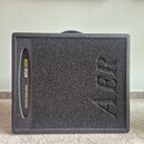 Černý basový kombo zesilovač s texturovaným povrchem. Na přední straně je vlevo nápis electric bass amp amp one a vpravo velký reliéfní nápis AER. Po…