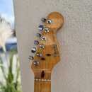 Fotografie zachycuje hlavu dřevěné elektrické kytary v přirozené barvě. Na hlavě je vidět logo Fender, nápis STRATOCASTER a text MADE IN USA. Pod log…