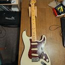 Elektrická kytara s bílým trupem, hnědým hnedem krku a hnedou želvovinovou pickguardem. Na hlavě krku je viditelné logo Fender. Kytara má tři snímače…