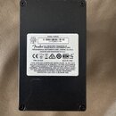 Fotografie zachycuje zadní stranu černého elektronického zařízení s viditelnými šrouby v rozích. Na přístroji je nalepený bílý štítek s nápisy 9VDC 2…