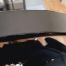 Detailní záběr na hranu černého lakovaného nástroje. Na povrchu laku jsou viditelné drobné škrábance a jedna výraznější oděrka v horní části hrany. Ž…