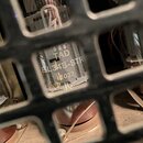 Fotografie zachycuje elektronovou lampu umístěnou za ochrannou mřížkou. Na skle lumpy je černý nápis TAD, EL34B-STR a číslo 1032. Povrch lumpy i okol…
